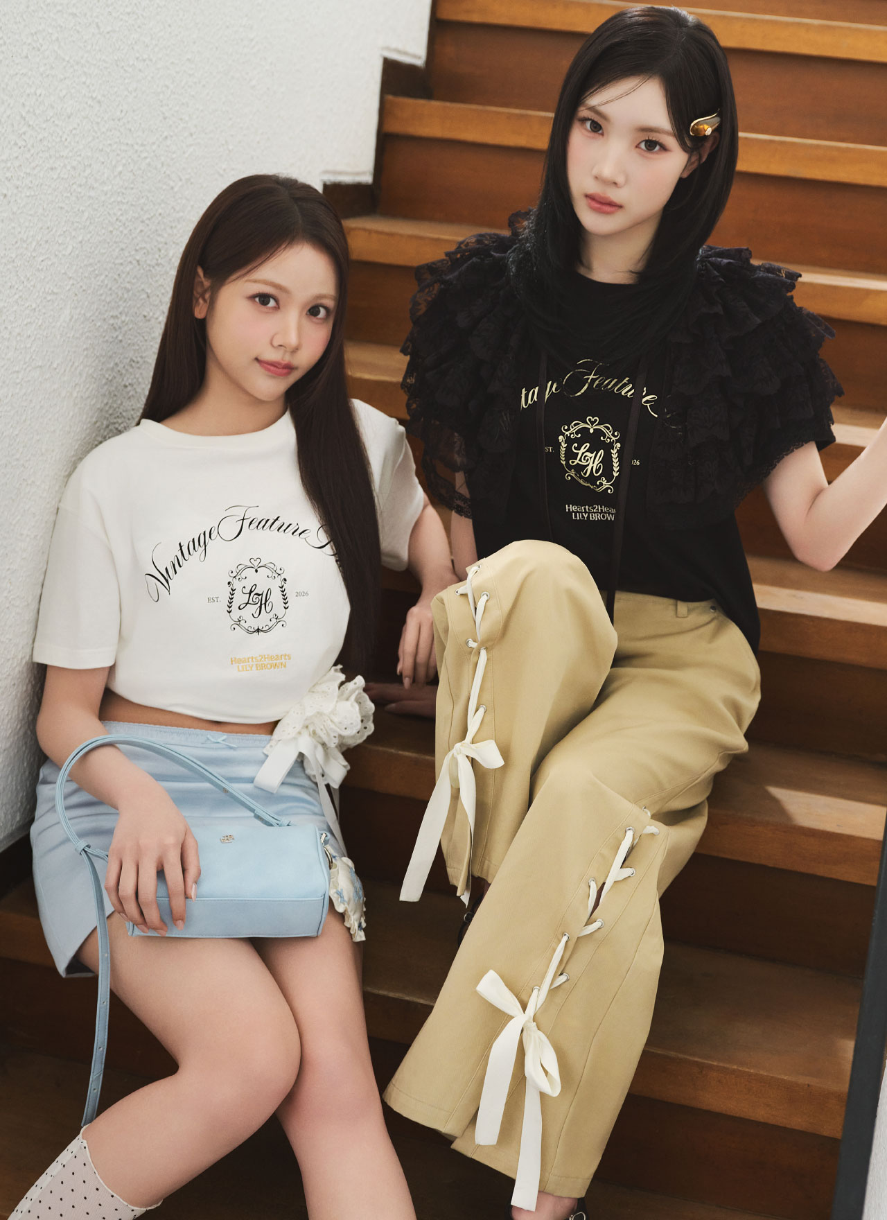 白いロゴTシャツの女性モデルと、黒いフリルトップスの女性モデルが階段に座る2ショットカット