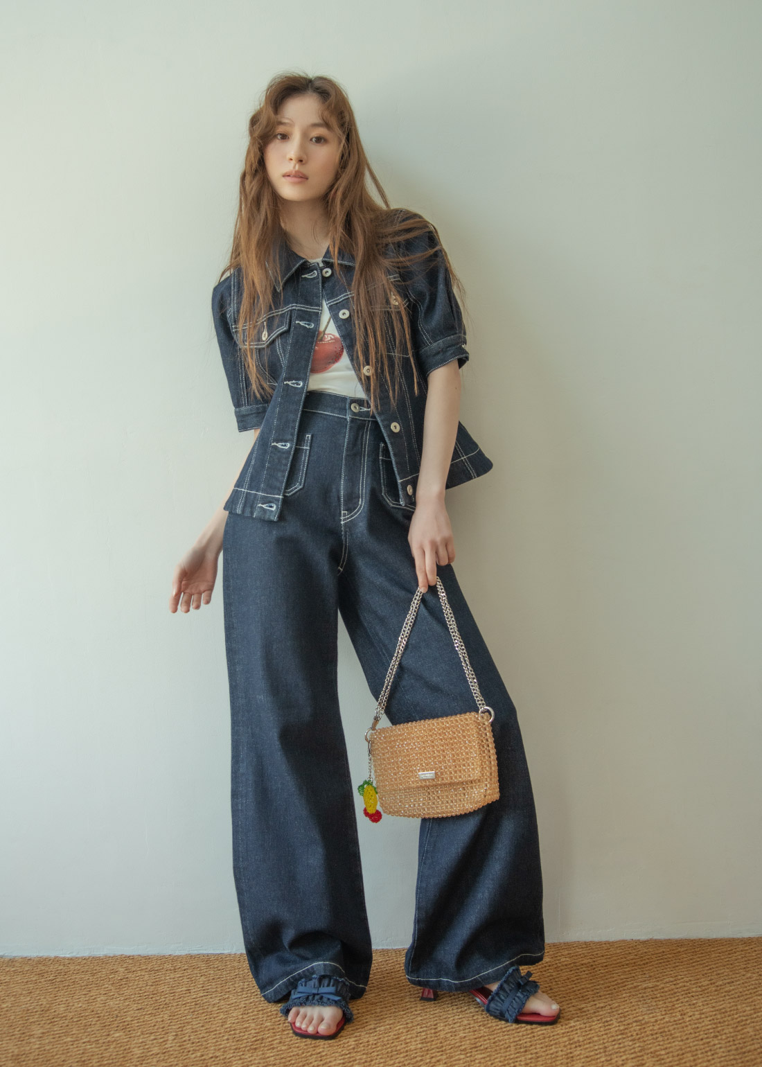 ネイビーのデニムジャケット・チェリーのイラストホワイトのトップス・ネイビーのデニムパンツ・デニムサンダル・ブラウンのバッグを着用した女性