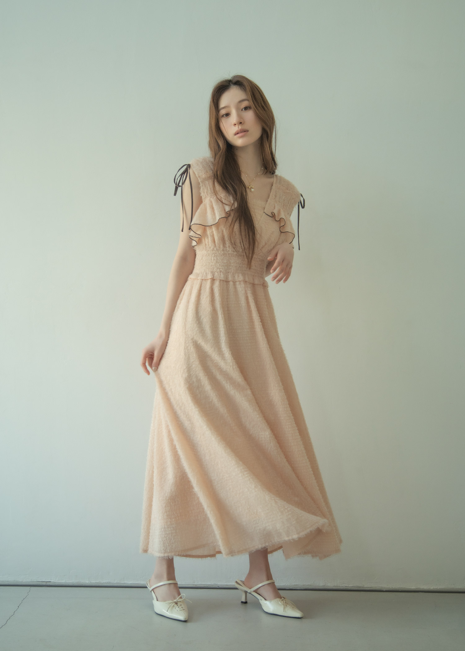 肩のリボンとフリルが特徴的な淡いピンクのロングワンピースに、白のパンプスを合わせた女性のコーディネート。